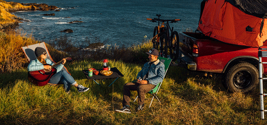Two people camping by the seaside, enjoying a meal and playing guitar next to a truck with a rooftop tent and bicycles. Ideal outdoor adventure setup with stunning ocean views.