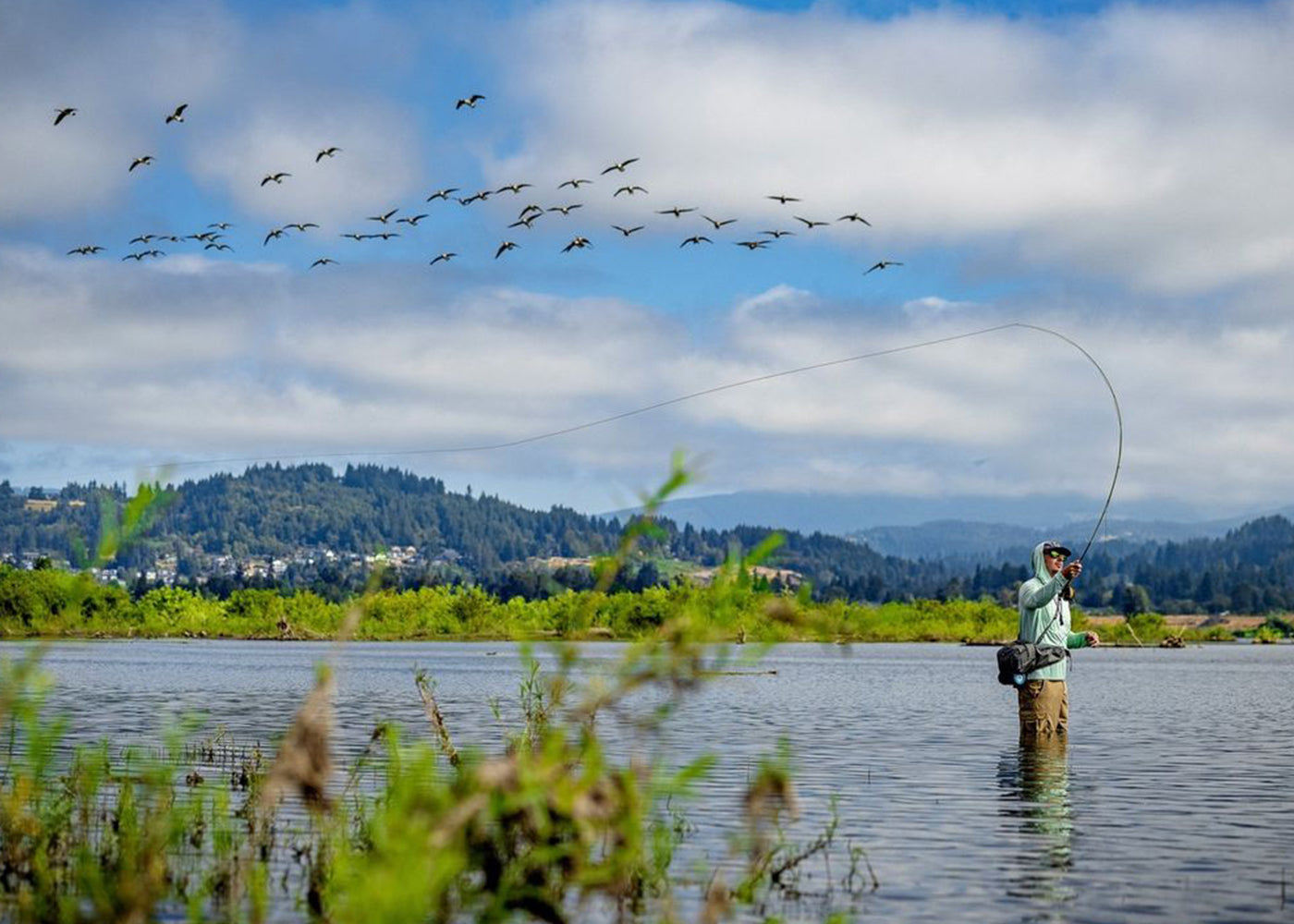 Fly fisherman using high-quality fishing gear while casting a line in a serene lake, with a flock of birds flying overhead. Scenic outdoor fishing experience with beautiful landscape views.