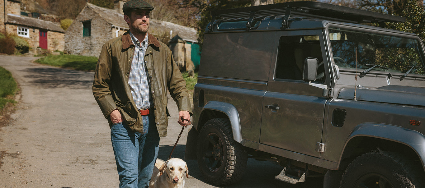 Man walking a dog next to a Land Rover, wearing a Barbour jacket and flat cap. Ideal for outdoor country style.
